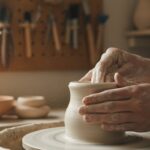 Macro photography of an artisan's hands shaping white kaolin clay on a pottery wheel.